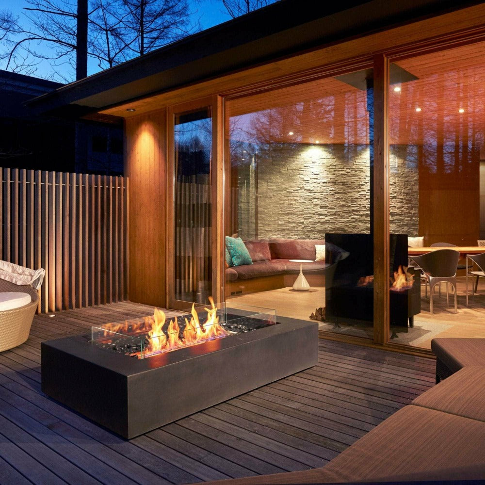 An outdoor fire pit with flames is set on a wooden deck, adjacent to a modern home with large windows revealing an interior seating area